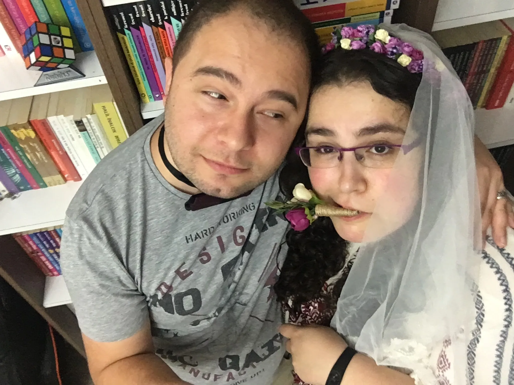 A man, Ömür Yanıkoğlu, wearing a bow tie and looking to a female, his wife Evrim Yanıkoğlu, who is wearing a veil and holding the groom's boutonniere in her mouth