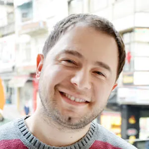 Ömür Yanıkoğlu. He is approximately 25 years old, has short hair, stubbly beard, wearing a gray red sweater and smiling.